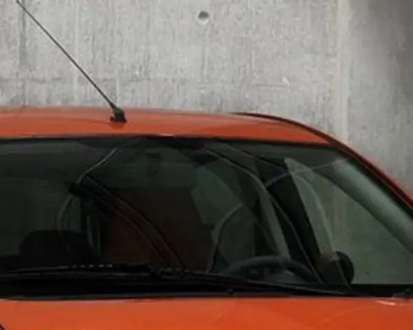 Pomarańczowy samochód z przyciemnianymi szybami z przodu, z antenką na dachu, stoi przy betonowej ścianie.
