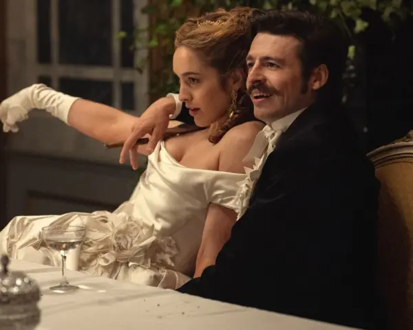 Elegancka para przy stole, zapowiedź nowych produkcji Netflix. Kobieta w białej sukni z rękawiczkami, mężczyzna w czarnym fraku.