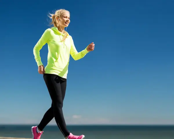 Uśmiechnięta blondynka w neonowej bluzie i czarnych legginsach uprawia power walking nad morzem.
