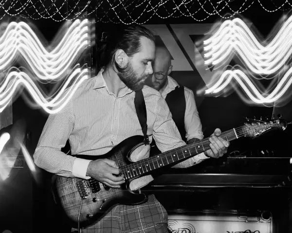 Muzyk gra na gitarze elektrycznej. W tle widać drugiego muzyka i falujące światła, tworzące efekt ekspozycji.