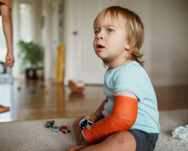 Kleiner Junge mit einem orangefarbenen Gips am Arm, der nachdenklich auf den Boden schaut. Er hat einen **kind arm gebrochen**.