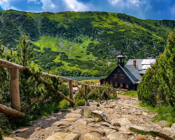 Kamienna ścieżka prowadzi do schroniska Samotnia Śnieżka nad jeziorem, otoczonego zielonymi zboczami gór.