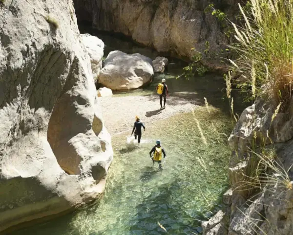 Trois personnes en combinaison font du canyoning dans une rivière aux eaux claires, entourées de falaises rocheuses.