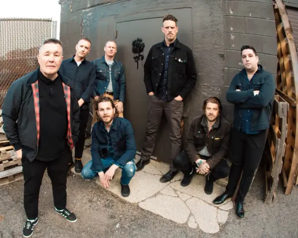 Les membres de Dropkick Murphys posent devant un mur sombre, certains accroupis, d'autres debout, prêts pour leur prochain concert.