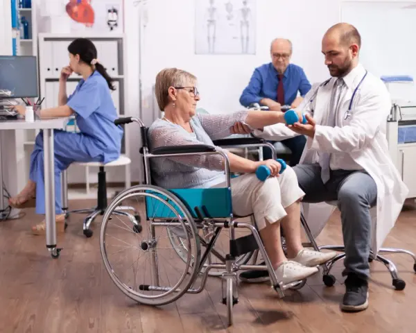 Rehabilitacja po uszkodzeniu móżdżku: pacjentka w wózku wykonuje ćwiczenia z hantlami pod okiem fizjoterapeuty.