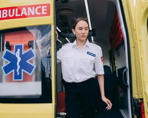 Młoda kobieta w białej koszuli z naszywką "PARAMEDICS" stoi przy otwartych drzwiach karetki. Czy kierowca karetki musi być ratownikiem?