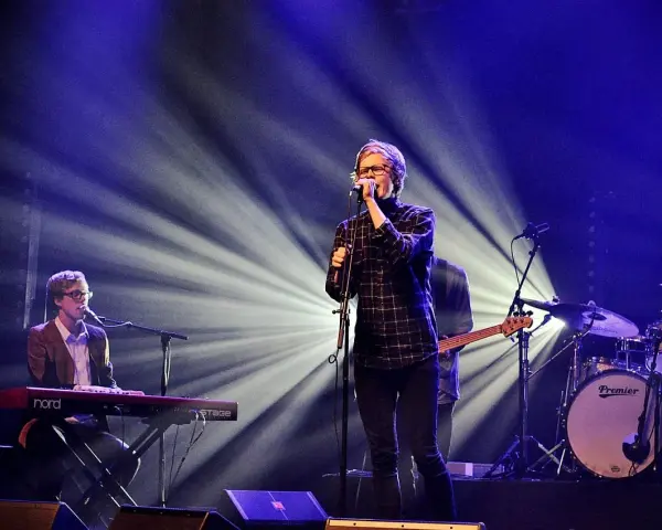 Zespół na scenie, wokalista śpiewa do mikrofonu, klawiszowiec gra na keyboardzie, a perkusista wybija rytm. Wspaniałe tło muzyczne.