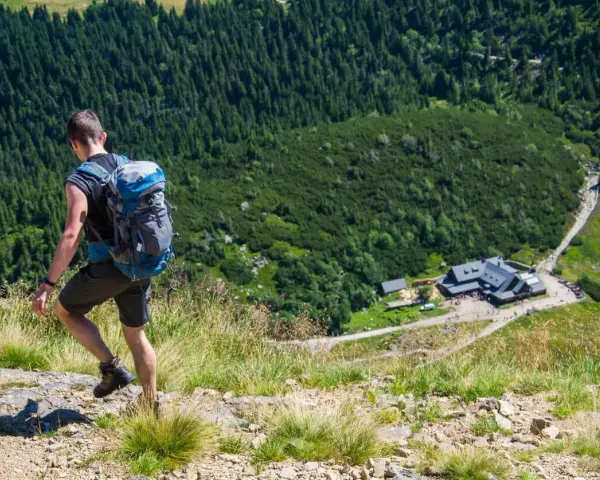 Turysta z plecakiem schodzi po kamienistej ścieżce w Karkonoszach. W dole widoczny schronisko, jedna z wielu atrakcji górskich.