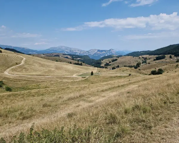 Ampia vallata con erba secca e un sentiero tortuoso che sale verso il passo fittanze. Montagne sullo sfondo sotto un cielo azzurro con nuvole bianche.