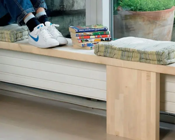 En person sitter på en bänk med kuddar och böcker. Under bänken finns ett element som är inbyggt i möbeln.