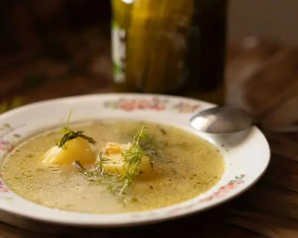 Aromatyczna keto zupa ogórkowa z ziemniakami i koperkiem, podana na talerzu z kwiatowym wzorem. Obok stoi słoik ogórków.