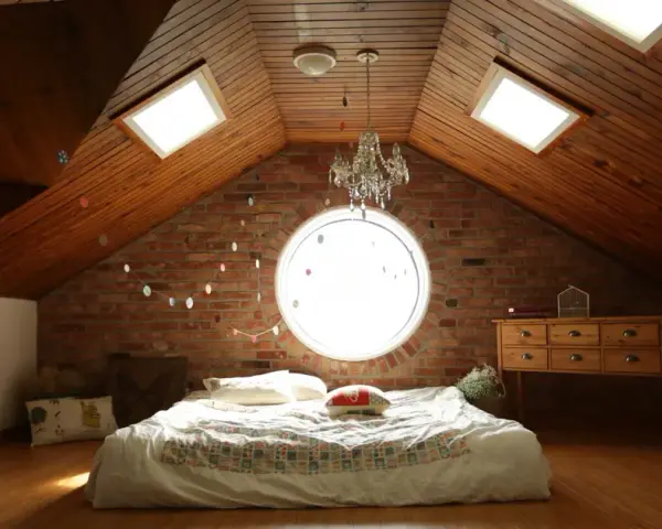 Camera puntata verso un letto in mansarda, con il soffitto in legno e la sua evidente pendenza tetto. Finestre sul tetto e un oblò circolare illuminano la stanza.