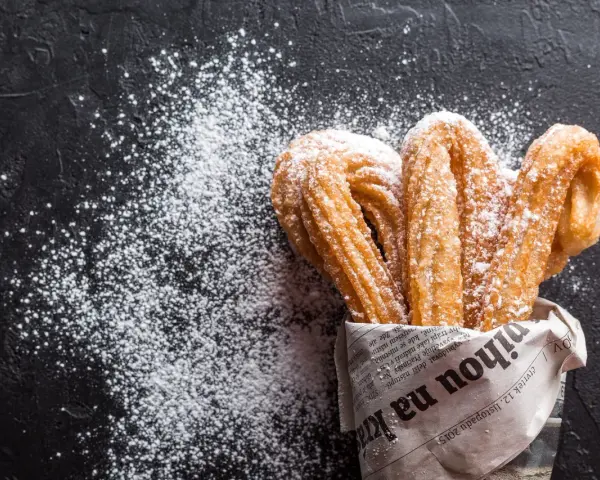Churros z czekoladą kontra pączki z lukrem? Te złociste churros, posypane cukrem pudrem, kuszą na czarnym tle.