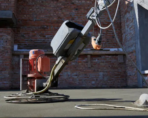 Robot do zacierania betonu pracuje na budowie. Robotnik w niebieskich spodniach kieruje maszyną, która wygładza świeżą posadzkę.