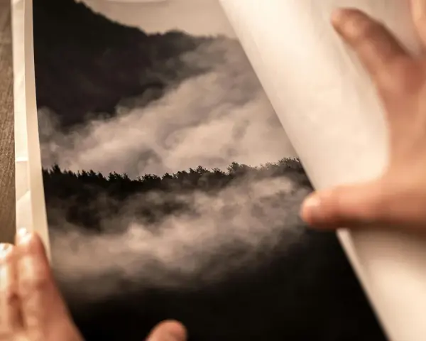 Dłonie otwierają album z fotografią górskiego lasu we mgle, wydrukowaną na papierze barytowym.