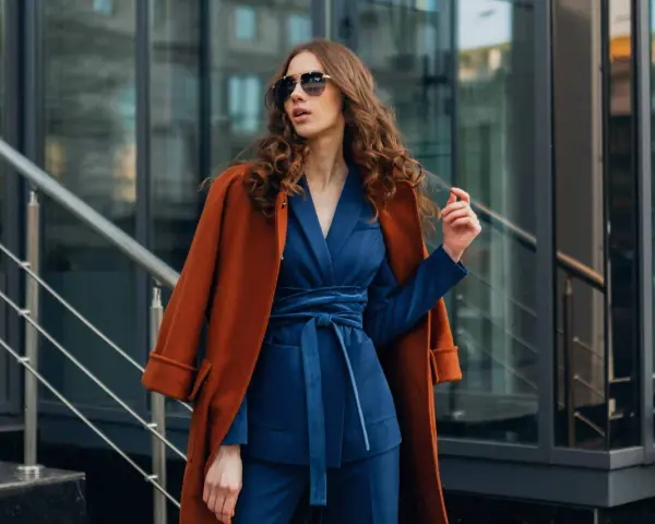 Une femme porte un gilet avec une robe bleu marine, un manteau orange sur les épaules, et des lunettes de soleil.