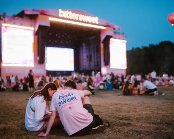 Para siedząca na trawie przed sceną festiwalową z napisem "bittersweet". W tle tłum ludzi i wielkie ekrany.