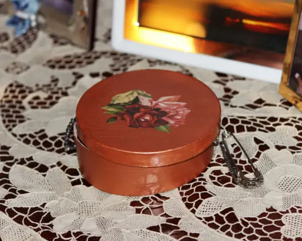 Okrągłe pudełko ozdobione techniką decoupage z motywem róż. Obok broszka i ramka ze zdjęciem.