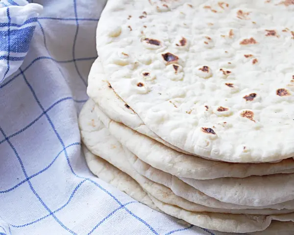 En hög med nybakade tortillabröd, perfekta för ditt nästa tortilla recept. Bredvid ligger en blåvitrutig kökshandduk.