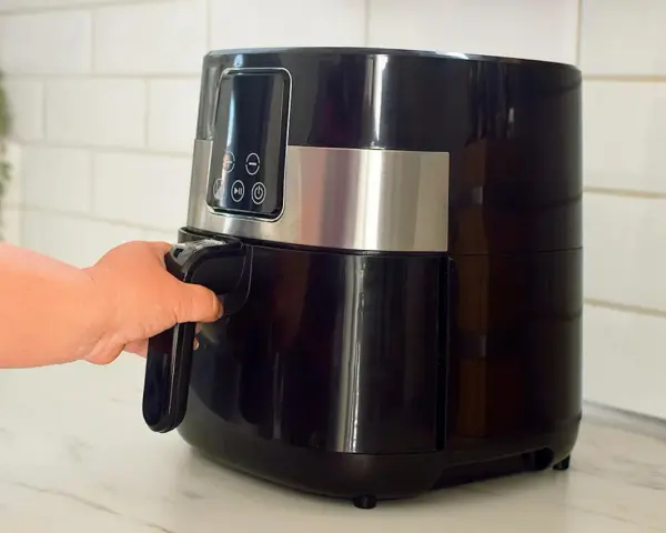 En hand drar ut korgen på en svart airfryer. Den här köksapparaten är lika praktisk som en diskmaskin för snabb matlagning.