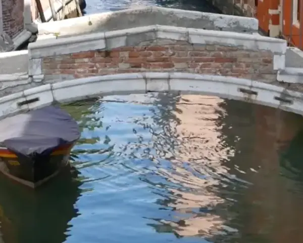Un piccolo ponte a Venezia, senza parapetto, sovrasta un canale dove una barca coperta attende.