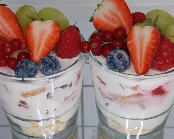 Zwei Gläser mit einem cremigen Dessert, garniert mit frischen Erdbeeren, Himbeeren, Blaubeeren, Johannisbeeren und Trauben. Ein köstliches Dessert im Glas.