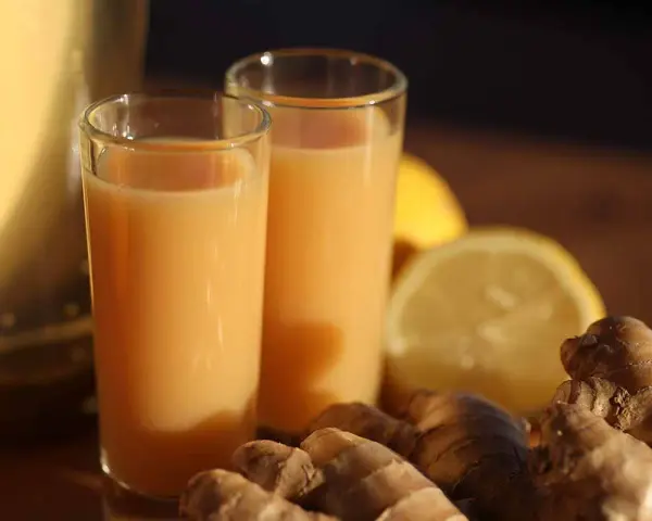 Zwei Gläser mit einem leuchtend orangen Ingwer Shot stehen neben frischem Ingwer und Zitronenscheiben.