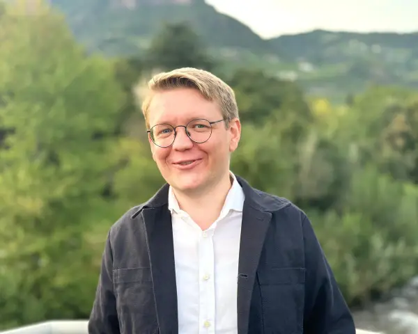 Ein Mann mit Brille lächelt in die Kamera. Er trägt ein weißes Hemd und eine dunkle Jacke. Im Hintergrund sind Bäume und Berge zu sehen. Er studiert Seelsorge.