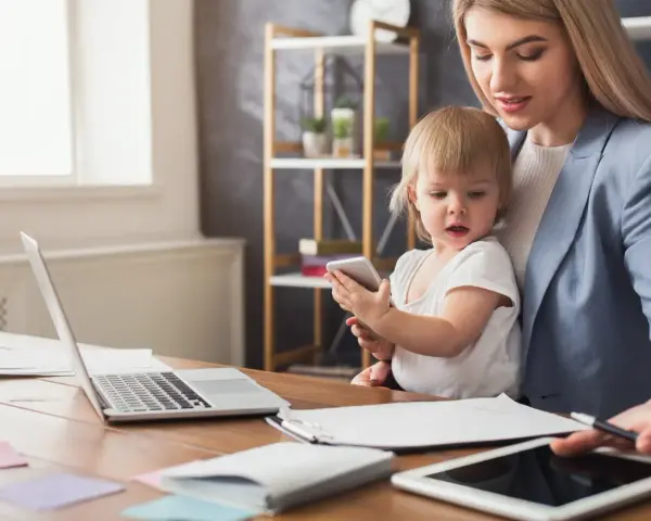 Pracująca mama z dzieckiem na kolanach, laptop i tablet na biurku. Czy na macierzyńskim można pracować? Tak, to możliwe!