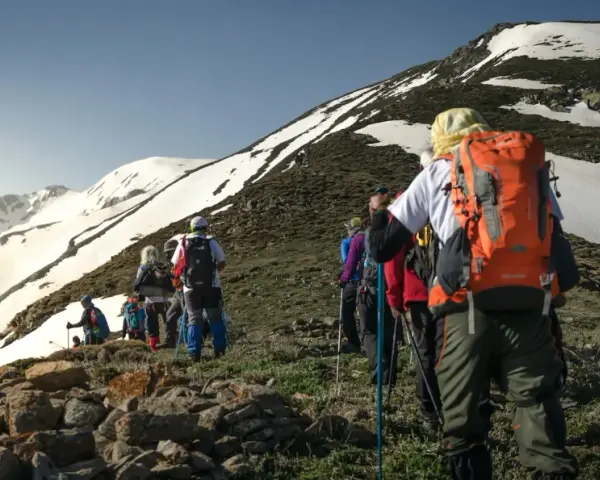 Un groupe de randonneurs progresse sur un sentier rocailleux, le soleil éclairant les montagnes enneigées. Ils semblent déterminés à savoir son altitude.