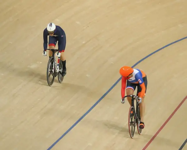 Two cyclists compete in a track sprint. One rider in a blue and white uniform leads, while another in orange and blue follows closely.