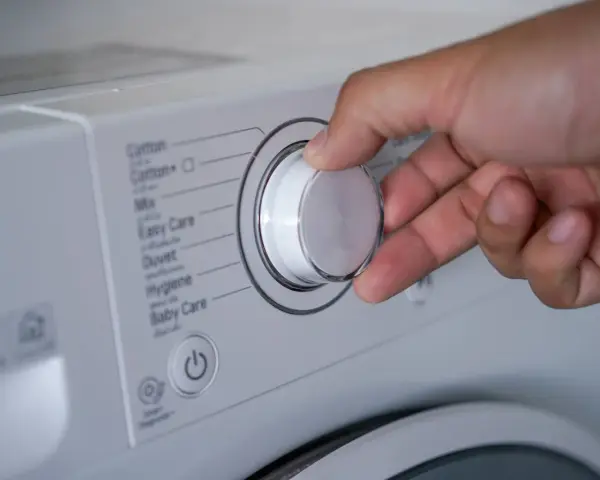 Mano ajustando el selector de la lavadora para saber como lavar un anorak. Se ven programas como "Cotton", "Mix", "Duvet".