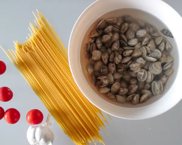 Składniki na pyszny makaron z małżami: spaghetti, pomidorki, czosnek i świeże małże w misce.