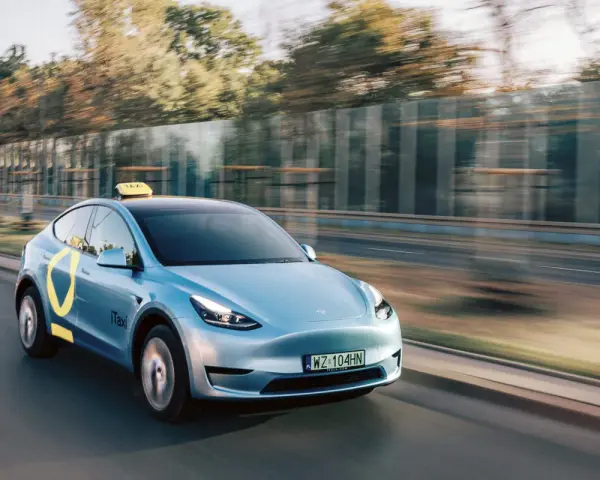 Niebieski Tesla Model Y z żółtym znakiem taxi na dachu i logo iTaxi na drzwiach, spełniający wymagania samochodu dla tej usługi.