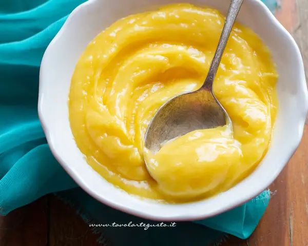 Una ciotola bianca piena di vellutata crema al limone, con un cucchiaino che ne raccoglie una porzione.