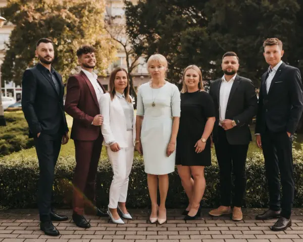 Grupa sześciu profesjonalistów w eleganckich strojach stoi przed budynkiem i zielenią.