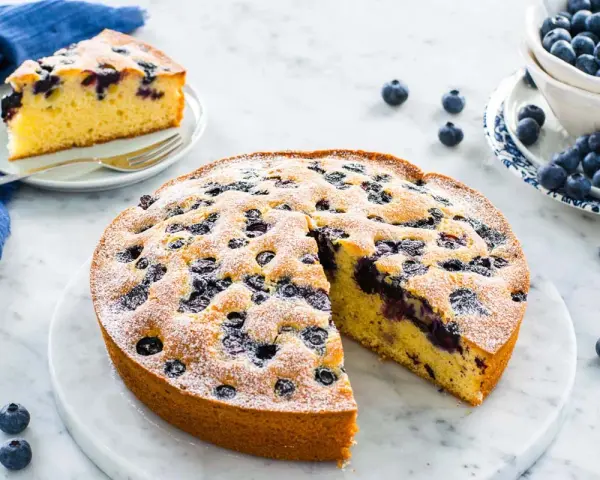 Deliziosa torta allo yogurt con mirtilli, spolverata di zucchero a velo. Una fetta è già servita su un piattino.