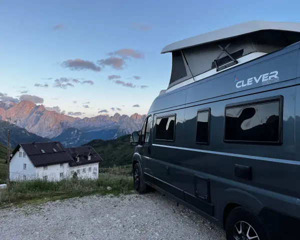 En mörkgrå husbil med pop-up tak, märkt "Clever vans", står parkerad vid en bergig stuga med Alperna i bakgrunden.