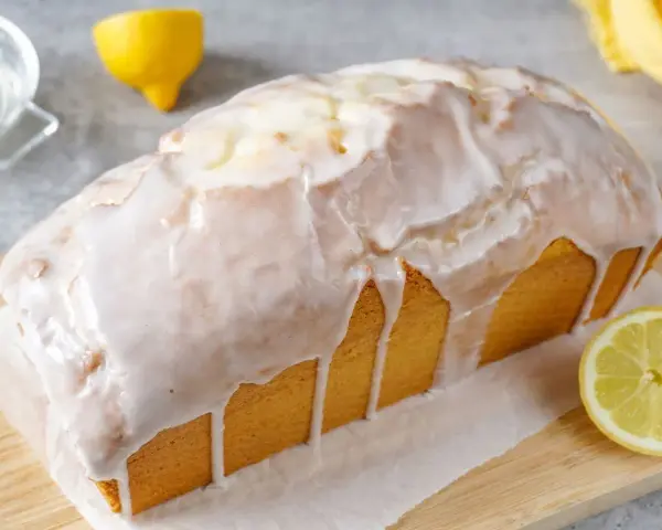 Saftiger zitronenkuchen in Kastenform, überzogen mit süßem Zuckerguss. Ein Zitronenstück liegt daneben.