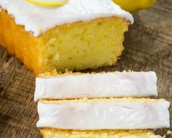 Fetta di plumcake al limone con glassa bianca e una fetta di limone sopra, su un tagliere di legno.