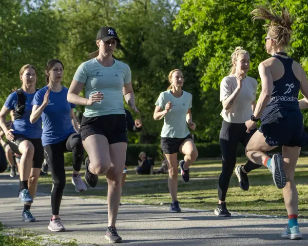 En grupp kvinnor tränar löpning utomhus. De springer i ett parkliknande område, redo att springa 1 km tid.