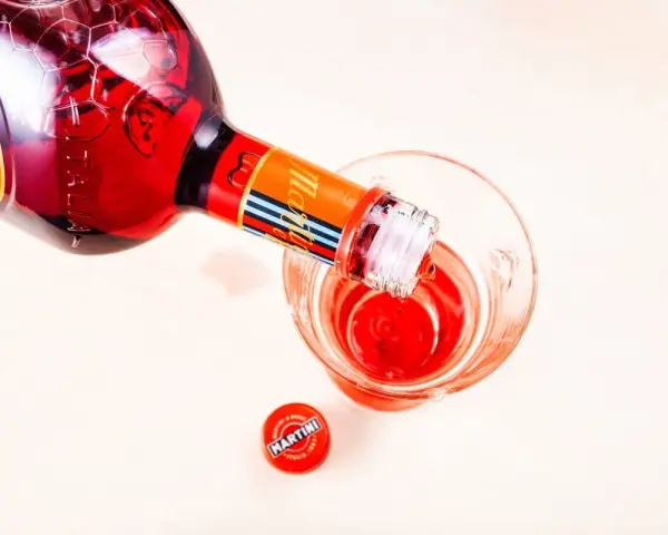 Czerwony Martini z sokiem pomarańczowym nalewany do kieliszka. Obok leży czerwona zakrętka z logo Martini.