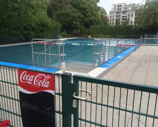 Erfrischung im Monbijou Bad: Wasser 21°C. Menschen schwimmen im blauen Becken, umgeben von Bäumen und einem modernen Gebäude.