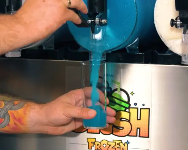 Ein Arm mit einem Ketten-Tattoo füllt ein Glas mit blauem Slush Eis aus einem Automaten.