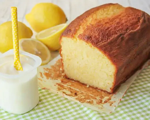 Bizcocho de yogur casero, dorado y esponjoso, acompañado de limones frescos y un vasito de yogur.