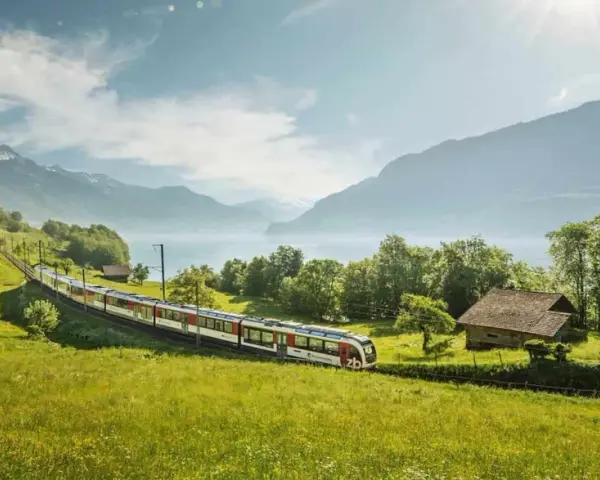 A train travels through a lush green valley, with a lake and mountains in the background. This scenic route from Lucerne to Montreux offers breathtaking views.