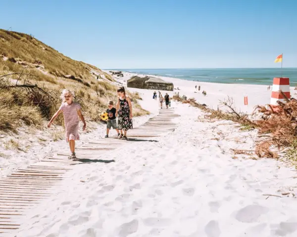 Familj promenerar på en strand med vit sand i Danmark. En livräddartorn och bunkrar syns i bakgrunden.