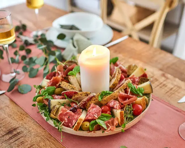 Antipasti selber machen: Eine festliche Platte mit Wurst, Käse, Brot, Oliven und Tomaten, umgeben von grünen Zweigen, mit einer Kerze in der Mitte.