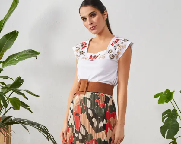 Mujer con top blanco bordado y falda plisada de estampado abstracto. Un cinturón marrón ancho realza la cintura.