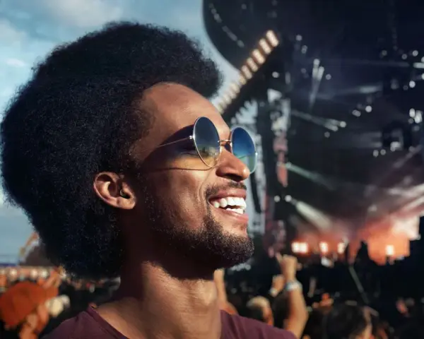 Homme souriant à un concert, portant des lunettes de soleil et un bouchon d'oreille pour protéger son audition.